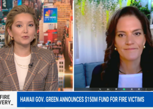 Two women speak on a news broadcast about Hawaii’s $150M fund for fire victims, with cityscape background.